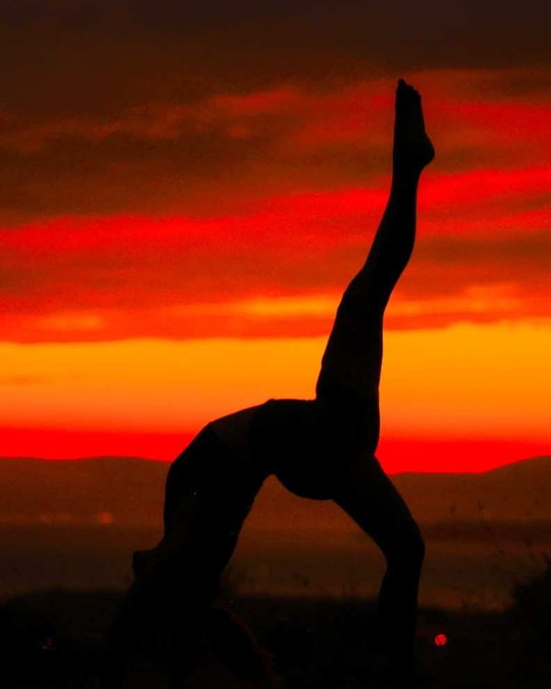 Silhouette of a one-legged backbend on a hillside beneath a red and orange twilight sky